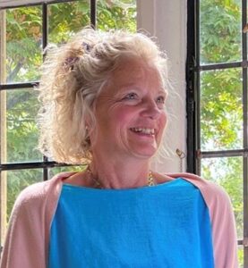 A woman with blond hair is standing in front of a window smiling. She is wearing a blue top and a pale pink cardigan. Outside it is sunny and there are trees in leaf.