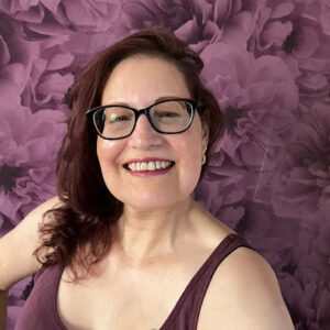 A woman with long dark wavy hair and glasses smiles at the camera. She is wearing a purple vest top. Behind her is a background of large purple flowers.