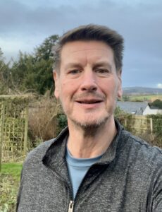 A man with short hair, a short beard and moustache stands in a garden. He is wearing a grey zip-up top. Behind him is a fence, a house and open countryside.
