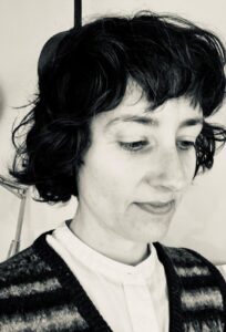 A woman with short dark curly hair smiles gently as she looks down. She is wearing a white blouse and woollen cardigan.