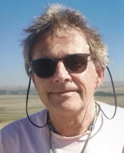 A man with short, windswept hair and sunglasses stands on a beach smiling.